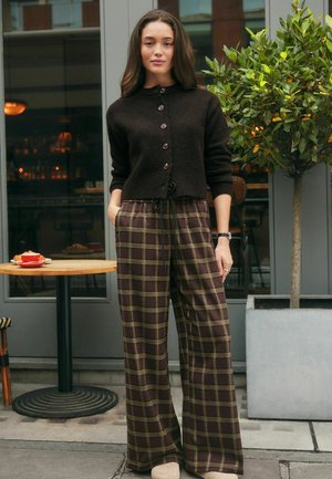 Hnedý pletený kardigán s gombíkmi, skombinovaný so širokými károvanými nohavicami s hnedými a zelenými štvorcami. Svetlé topánky dotvárajú outfit.