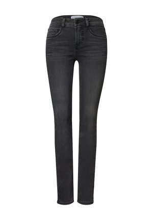 Person iført en brun hundetandsjakke over en hvid skjorte med den ene hånd i jakkelommen og sorte jeans.