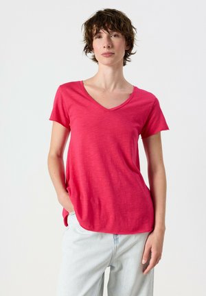 Jeune personne aux cheveux bruns courts portant un t-shirt rose vif à col en V et un jean bleu clair, debout avec une main dans la poche.