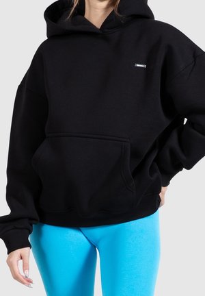 Person wearing a black oversized hoodie with a front pocket and blue leggings, standing with one hand on hip against a plain background.