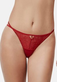 String en dentelle rouge avec motifs floraux, petit détail en bijou central, et fines bretelles. Tissu lisse avec une coupe minimaliste.