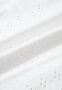 Tissu blanc avec une délicate broderie à jours formant des motifs géométriques et floraux, présenté en plis doux.