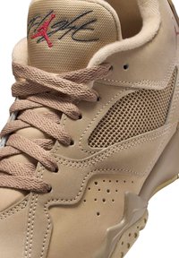 Beige sneaker with mesh side panels, padded tongue, and textured laces. Features a red logo and small perforations on the sole.