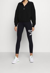Pull-over en polaire noire avec une fermeture éclair au quart, associé à un legging noir taille haute avec un grand logo blanc Reebok. Chaussures de sport blanches.