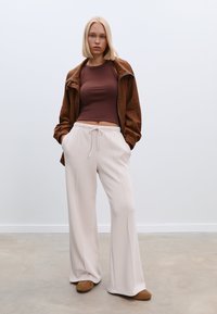 Braune Kunstlederjacke über einem enganliegenden braunen Oberteil, kombiniert mit weit geschnittenen, hellbeigen Hosen und braunen Slippern. Glatte Materialien, entspannte Passform.