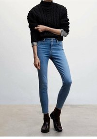 Suéter negro de punto trenzado con cuello alto, sobre una camiseta de manga larga a rayas. Jeans ajustados de mezclilla azul claro y calcetines marrones con mocasines negros.