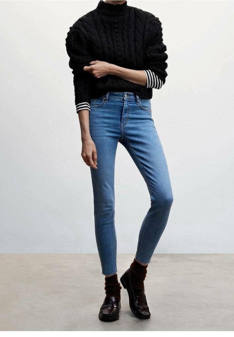 Suéter negro de punto trenzado con cuello alto, sobre una camiseta de manga larga a rayas. Jeans ajustados de mezclilla azul claro y calcetines marrones con mocasines negros.