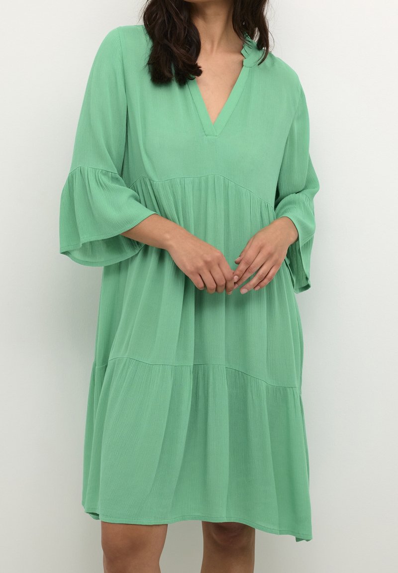 Femme portant une robe ample longueur genou vert menthe avec de larges manches évasées trois quarts et un col en V sur un fond clair uni.