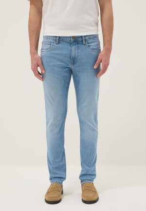 Hombre con vaqueros ajustados azul claro, camiseta blanca y mocasines de ante color canela, de pie frente a un fondo claro y liso.