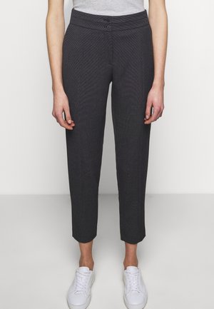 Persona che indossa pantaloni grigio scuro strutturati alla caviglia con vita a doppio bottone e sneaker bianche, in piedi davanti a uno sfondo semplice.