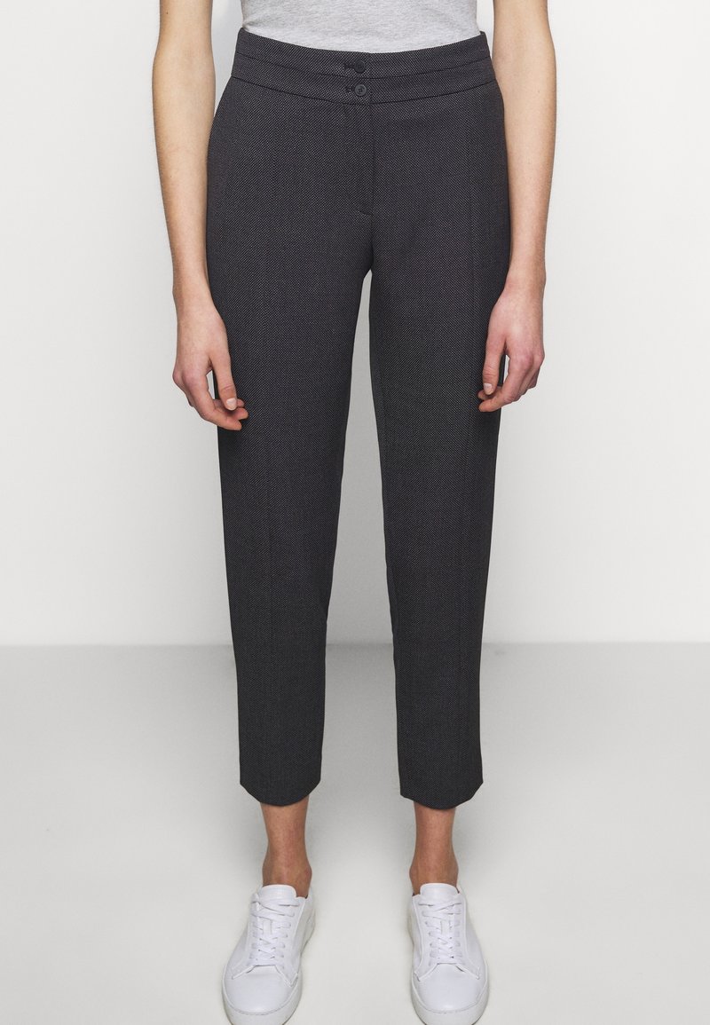 Personne portant un pantalon gris foncé texturé jusqu’à la cheville avec une ceinture à double bouton et des baskets blanches, debout devant un fond uni.