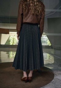 Chaqueta de cuero marrón, falda plisada gris oscuro y zapatos de tacón marrones. Texturas suaves con patrones sutiles, ambientado en un entorno interior.