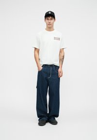 Witte t-shirt met korte mouwen en rode graphic; blauwe gestreepte wijde broek; zwarte pet; zwarte schoenen met een gestructureerd oppervlak.