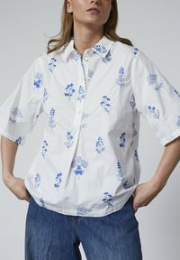 Hvid bomuldsbluse med korte ærmer, som har bl�å blomsterbroderi, knaplukning fortil og en afslappet pasform. Kombineret med denimbukser.