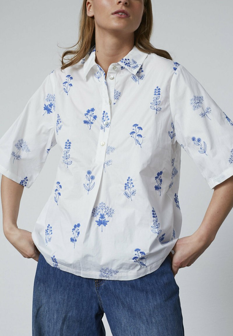 Hvid bomuldsbluse med korte ærmer, som har blå blomsterbroderi, knaplukning fortil og en afslappet pasform. Kombineret med denimbukser.