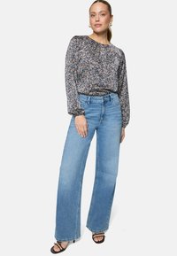 Femme portant un chemisier à manches longues à motifs floraux rentré dans un jean taille haute large et bleu clair, avec des sandales noires à talons et lanières.