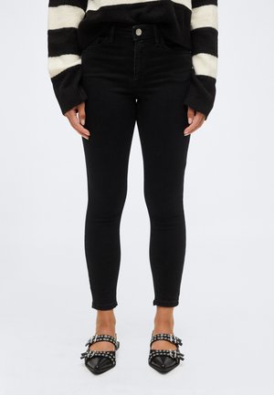 Femeie purtând blugi skinny negri cu talie înaltă, pulover în dungi alb-negru și pantofi negri ascuțiți cu catarame și ținte.