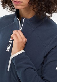 Marinefarbener Quarter-Zip-Pullover aus strukturiertem Material, mit weißen Reißverschlüssen und Logoakzenten, strukturiertem Kragen und enganliegenden Ärmeln.