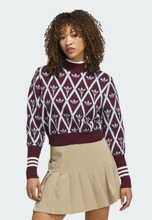 Woman with curly hair wearing a maroon patterned sweater and beige pleated skirt, standing against a plain light background.