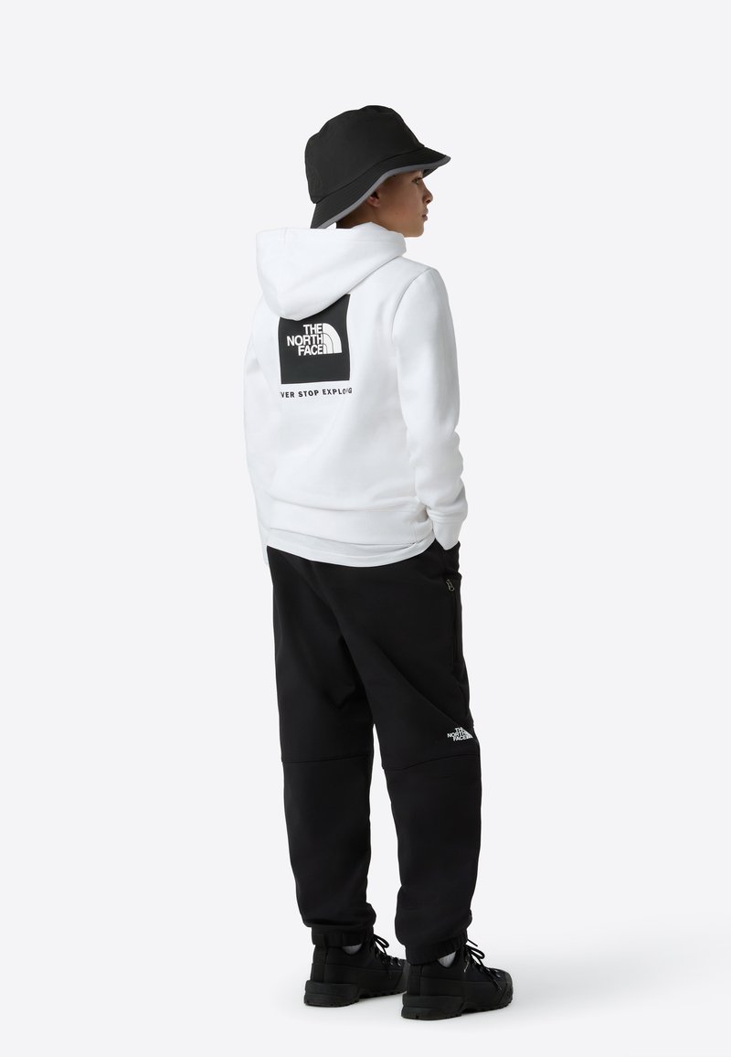 Hanorac alb cu un logo negru pe spate, purtat împreună cu pantaloni de trening negri. Modelul poartă o pălărie tip bucket neagră și încălțăminte cu talpă texturată.