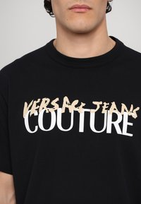 Juodas medvilninis marškinėlis su dideliu baltu užrašu ir smėlio spalvos nudėvėtu logotipu "VERSACE JEANS COUTURE" per krūtinę.