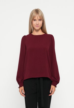 Blouse bordeaux à manches longues avec encolure ronde, tissu texturé, manches bouffantes et poignets élastiques ; assortie à un pantalon noir.