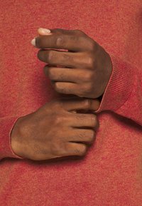 Pull en maille de couleur rougeâtre, doté de poignets côtelés, les mains placées au niveau des manches, révélant la texture et une nuance bicolore.