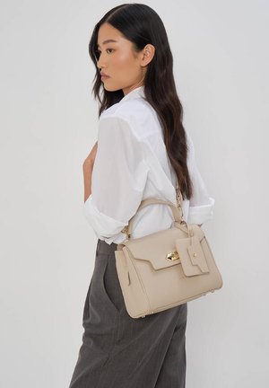 Donna con lunghi capelli scuri, indossa una camicia bianca e pantaloni grigi, porta una borsa beige con fibbia dorata e una pochette attaccata sulla spalla.