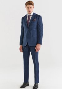 Granatowy w kratkę garnitur z marynarką na dwa guziki, pasującymi spodniami, białą koszulą i wzorzystym krawatem. Akcentująca chusteczka w kieszeni dodaje detali.