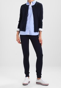 Marinblå cardigan, ljusblå skjorta, marinblå leggings och vita sneakers med röda och blå ränder. Slätt tyg, figurnära design, avslappnad outfit.