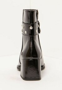 Botines negros con acabado brillante, tiras con tachuelas plateadas y tacón ancho. Presentan un cierre de cremallera y una textura suave.