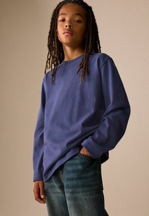 Ung person med lange dreadlocks iført en blå langærmet skjorte og jeans, poserer med den ene hånd i lommen mod en ensfarvet baggrund.
