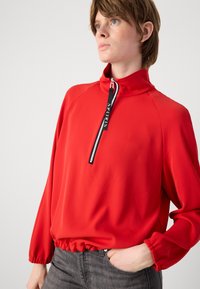 Rode ritssluiting pullover met hoge kraag, elastische zoom en lange mouwen. Voorzien van een zwarte trekband met de tekst "SPORTS". Zacht, glad materiaal.