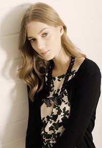Cardigan nero sopra una maglietta bianca con motivi floreali verdi. Una collana nera spessa e degli occhiali da sole poggiano sopra la maglietta. Capelli morbidi e mossi.