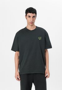 T-shirt noir surdimensionné avec des manches courtes, encolure ronde et un petit motif vert sur la poitrine. Fabriqué en tissu doux et texturé.