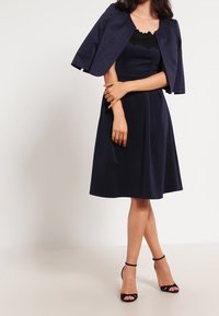 Abito blu navy con corpino aderente e gonna a campana, dotato di scollo in pizzo. Accompagnato da un giubbotto cropped blu navy con texture.