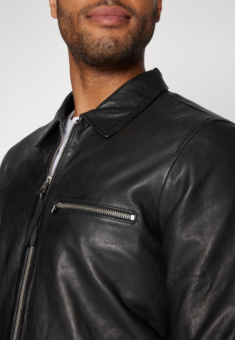 Schwarze Lederjacke mit glatter Textur, spitzem Kragen und einer Reißverschlusstasche auf der Brust. Silbere Details betonen das Design.