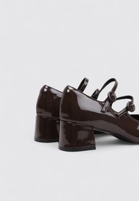 Zapatos de tacón de charol marrón con punta redonda, correa en el tobillo y un tacón cuadrado de 3 pulgadas que presenta pequeños acentos circulares.