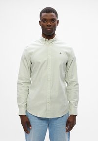 Ανοιχτό π ράσινο πουκάμισο με ρίγες, με μακριά μανίκια, pointed collar και μικρό κεντημένο λογότυπο στο αριστερό μέρος της θηλής. Φορεμένο με ανοιχτό μπλε τζιν.