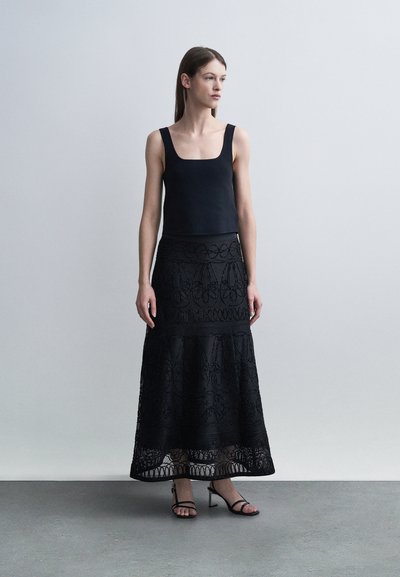 Femme debout portant un haut noir sans manches, une jupe longue noire brodée et des sandales à talons à lanières noires, devant un fond gris.