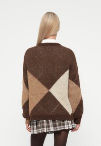 Vrouw met lang blond haar, gekleed in een geometrisch patroon trui in bruin, beige en crème, en een geruite rok, gezien vanuit de achterkant.