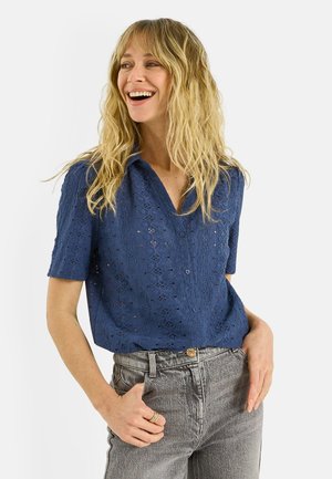 Femme blonde souriante, portant une chemise à manches courtes bleu marine avec des boutons ajourés et un jean taille haute gris, les mains dans les poches.