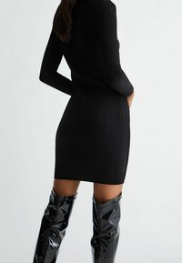 Abito lungo a maniche lunghe nero a costine con una silhouette aderente, abbinato a stivali neri lucidi fino al ginocchio, che mostrano una texture liscia e elegante.