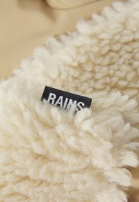 Kreminiu spalvos tekstūruotas vilnos audinys su pūkuotu, minkštu paviršiumi; turi juodą etiketę su balta užrašu "RAINS."