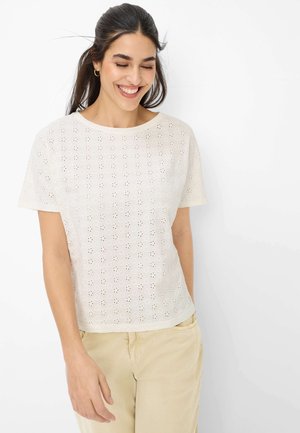 Smilende kvinde med langt mørkt hår iført en hvid øjenlet kortærmet top og beige bukser, stående foran en ensfarvet hvid baggrund.