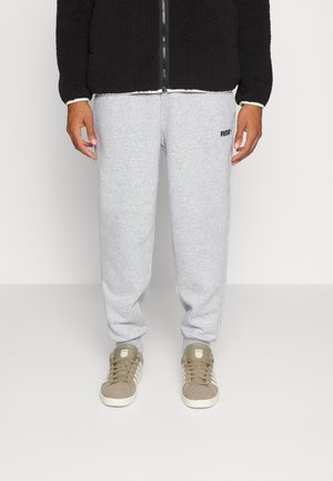 Personne portant un pantalon de survêtement Puma gris clair, des baskets beiges et une veste zippée noire, debout sur un fond clair uni.