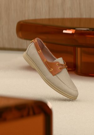 Mocassin beige clair avec garniture et lacets en cuir marron clair, présenté sur un tapis crème avec une table translucide marron en arrière-plan.