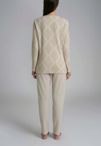 Donna in piedi a piedi nudi, girata di spalle, indossa un completo lounge beige con maniche lunghe e un motivo a diamante sulla parte superiore, pantaloni semplici, contro uno sfondo grigio.