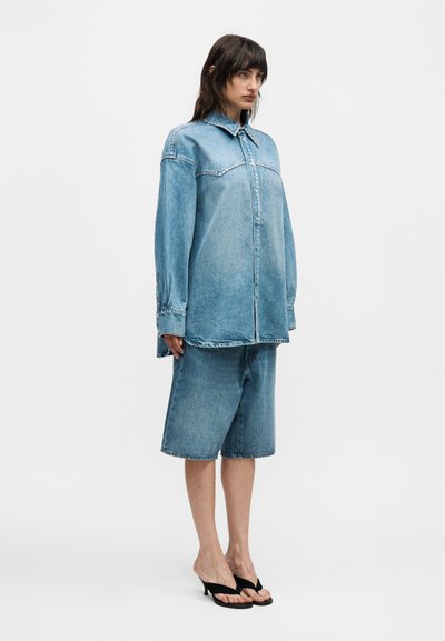 Γυναίκα στέκεται με φαρδιά ανοιχτό μπλε τζιν πουκάμισο και τζιν σορτς μέχρι τα γόνατα με μαύρα σανδάλια με τακούνι σε λευκό φόντο.