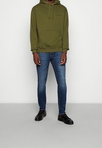 Olivově zelená mikina s kapucí, přední kapsou a šňůrkou, spárovaná s modrými slim-fit džíny a černými teniskami. Jednoduché barvy, neformální design.